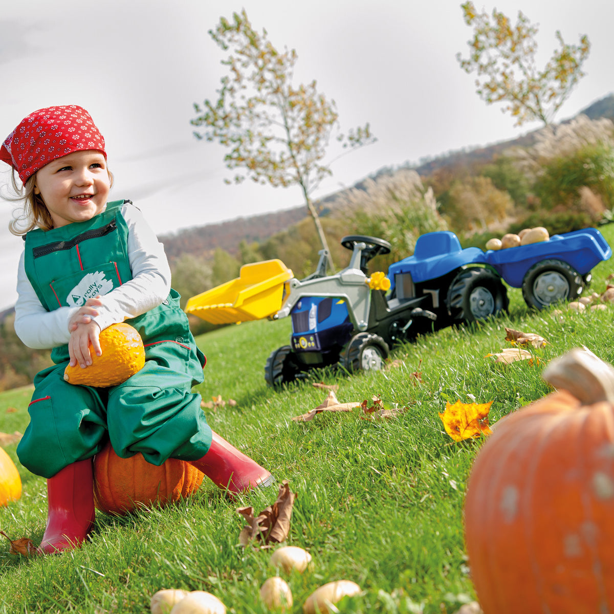 Outdoor lifestyle of the New Holland Kid Front Loader with Trailer with a little girl sitting on a pumpkin next to the tractor